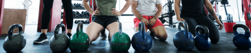 Three people in a weight room kneeling on a mat with various kettle bell weights in front of them.